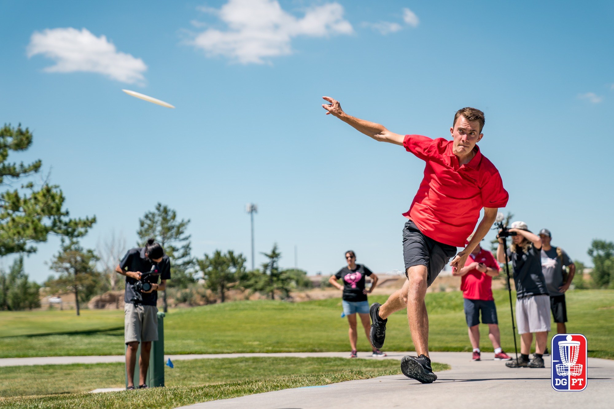 Will Schusterick releasing a drive at a professional disc golf event, disc in flight against a blue sky