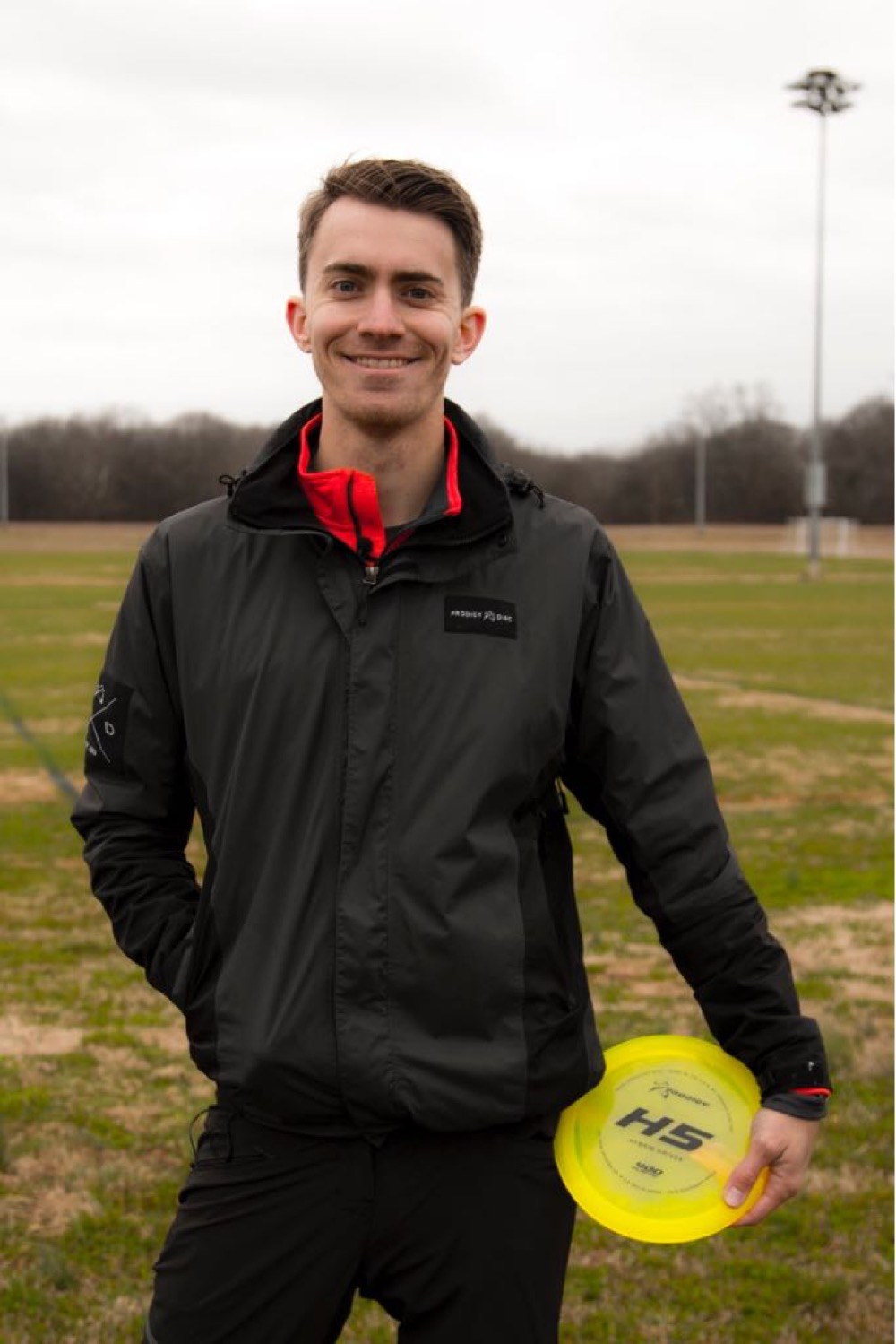 Will Schusterick in a Prodigy Disc jacket, holding an H5 driver on a course